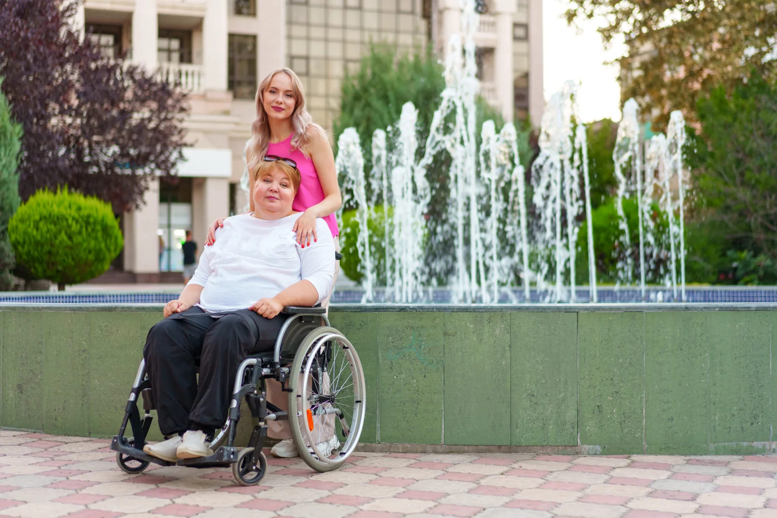 A woman using a wheelchair and a standing companion pause together beside a fountain in an outdoor plaza.