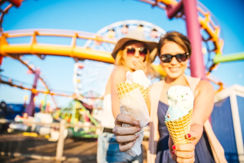Two women hold ice-cream cones with colorful amusement rides behind them.
