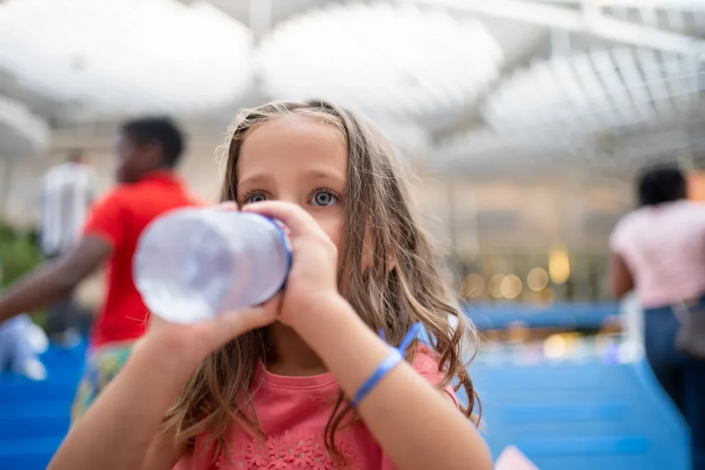 A young girl drinks from a water bottle in a busy covered public space.