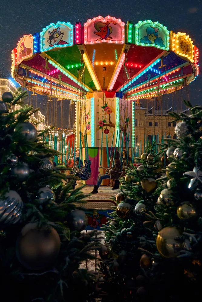 A brightly lit carousel stands between Christmas trees decorated with large shiny ornaments at night.