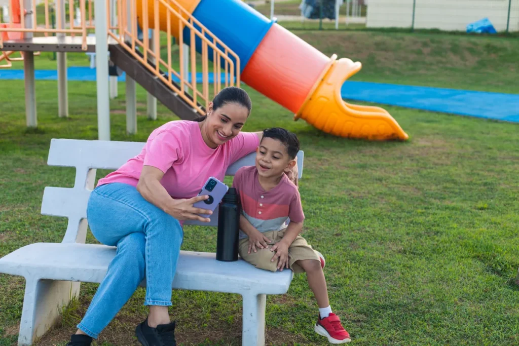 A woman and a boy sit together on a bench near colorful playground equipment.
