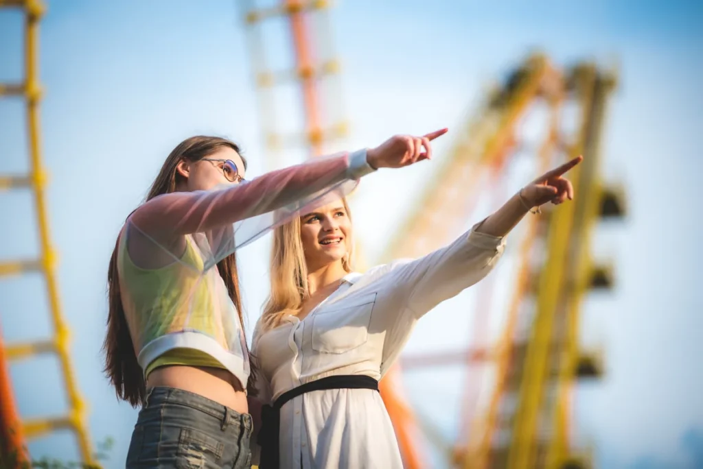 Two women point toward a ride with roller-coaster track behind them.