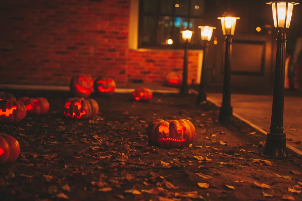 Carved pumpkins rest beside glowing lampposts on a leaf-covered path after dark.