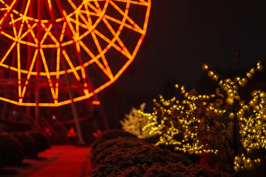 An orange wheel-shaped light display rises beside small trees strung with warm yellow lights at night.