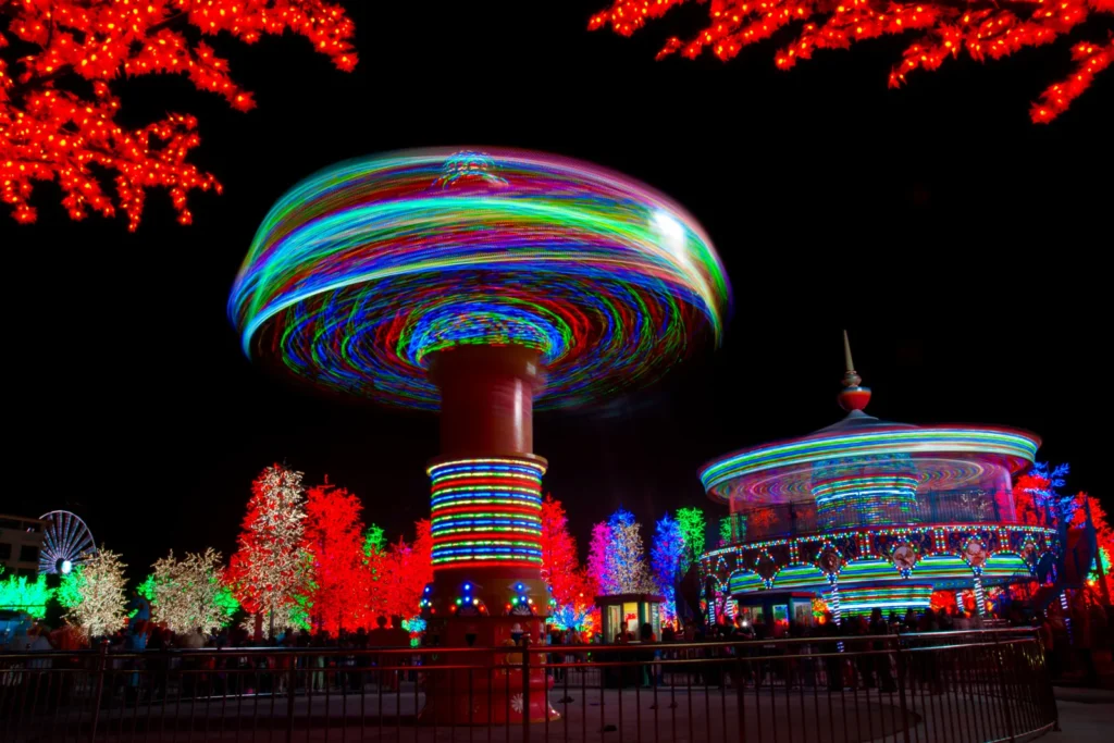 Colorful light trails circle two spinning amusement rides beside trees covered in orange, green, and white lights.