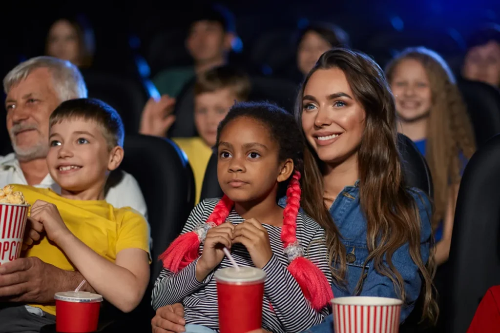 Adults and children watch a film in a dark theater with popcorn and drinks.