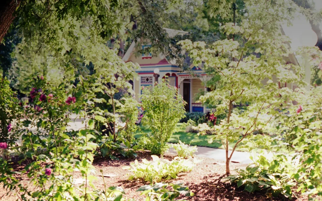 A historic building stands behind trees and a sunlit garden in Pioneer Village at Lagoon.