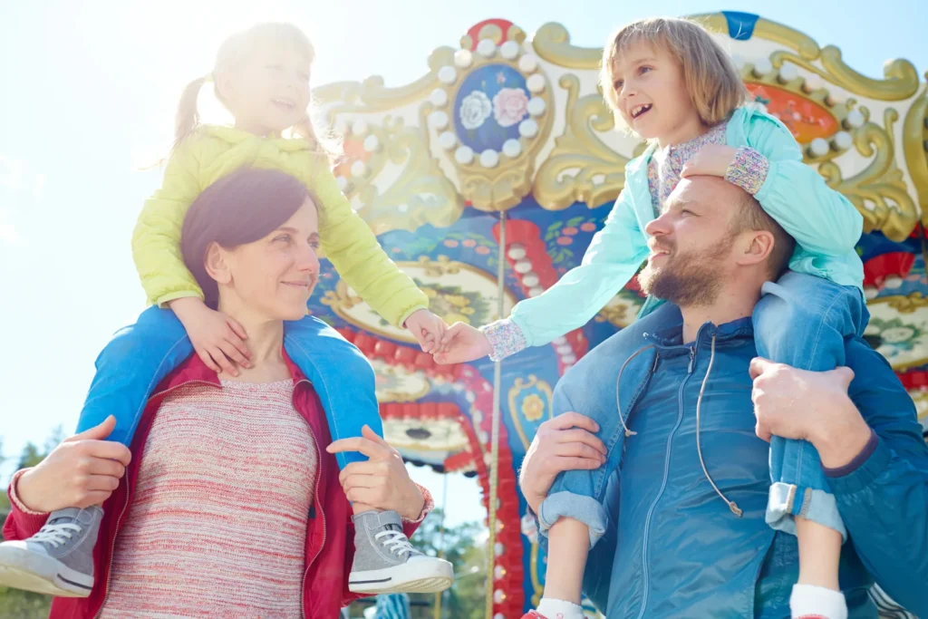 Two adults carry smiling children on their shoulders in front of a carousel.