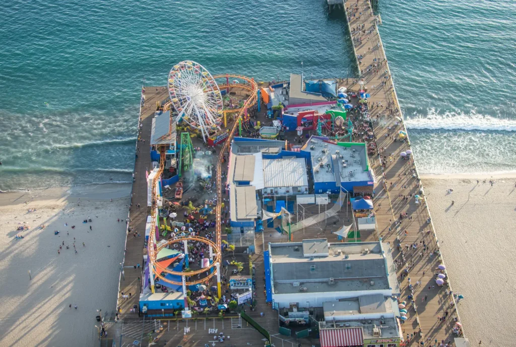 Aerial view of Santa Monica Pier showing a white Ferris wheel, colorful roller-coaster track, visitor walkways, beach, and ocean.