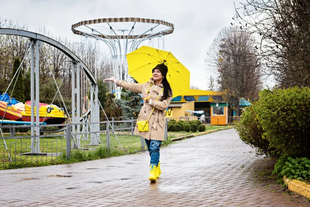 A woman holds a yellow umbrella beside amusement rides on a wet paved path.