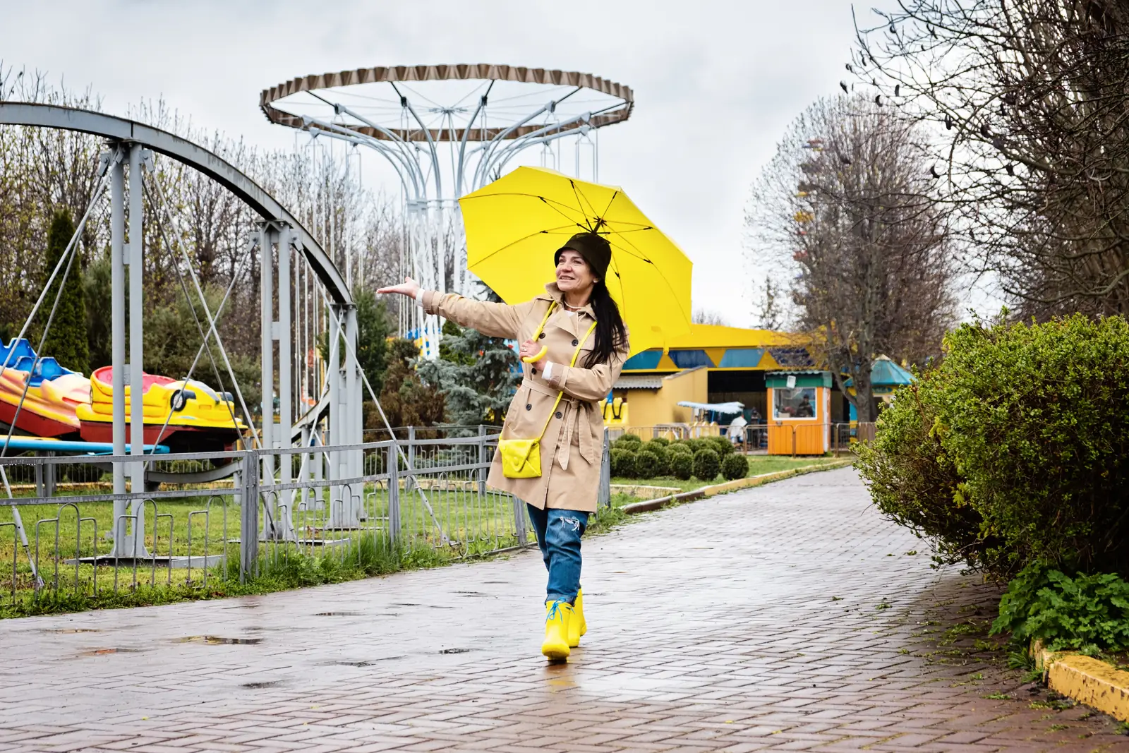 A woman holds a yellow umbrella beside amusement rides on a wet paved path.