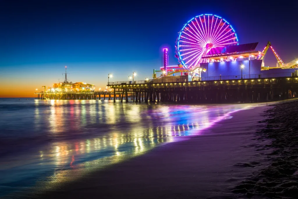 A glowing Ferris wheel rises above Santa Monica Pier as colored lights reflect on the wet beach.