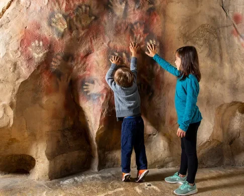 Two children match their hands to painted handprints in a model cave at the Smithsonian National Museum of Natural History.