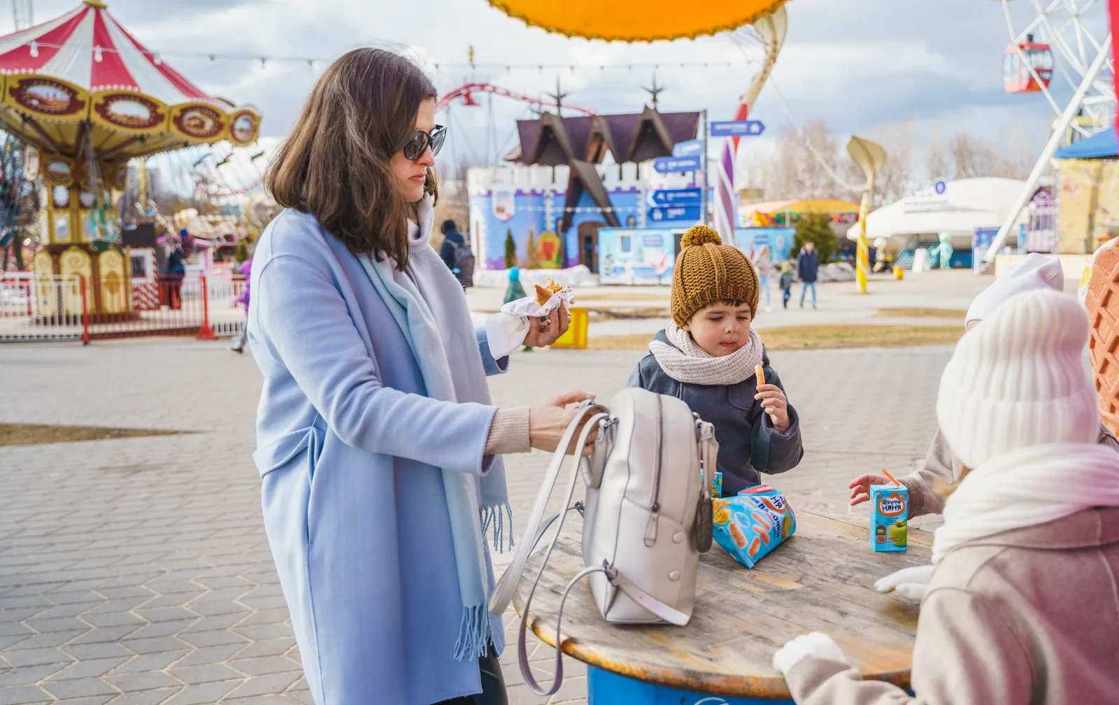 A woman in a pale blue coat and two children in knit hats share snacks at a round outdoor table with amusement rides behind them.