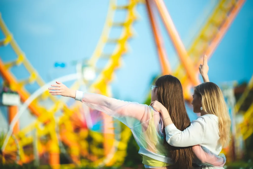 Two women stand together with arms outstretched in front of yellow roller-coaster track.