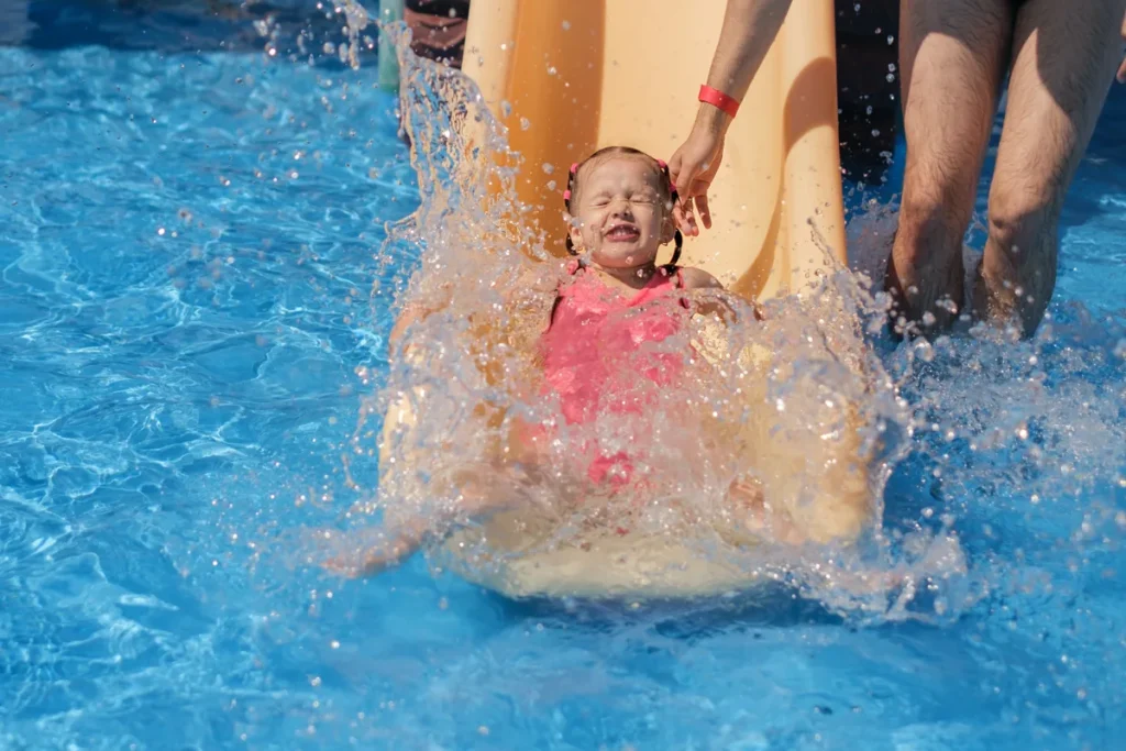 A young girl splashes into a pool from a small yellow water slide with an adult nearby.