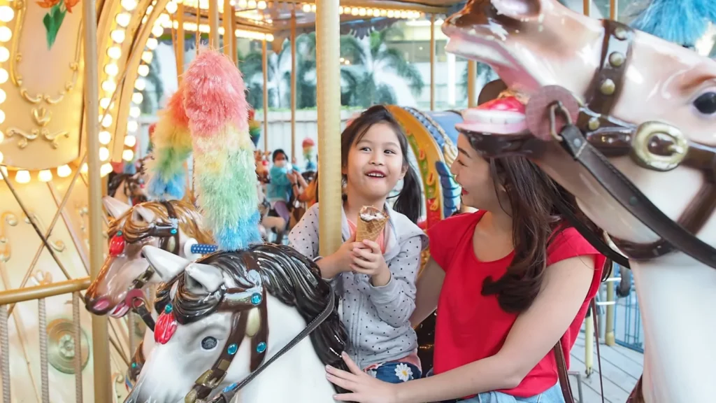 A child holding an ice-cream cone sits on a carousel horse beside an adult.